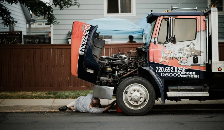 A mechanic works under a truck on a neighborhood street, highlighting vehicle maintenance.