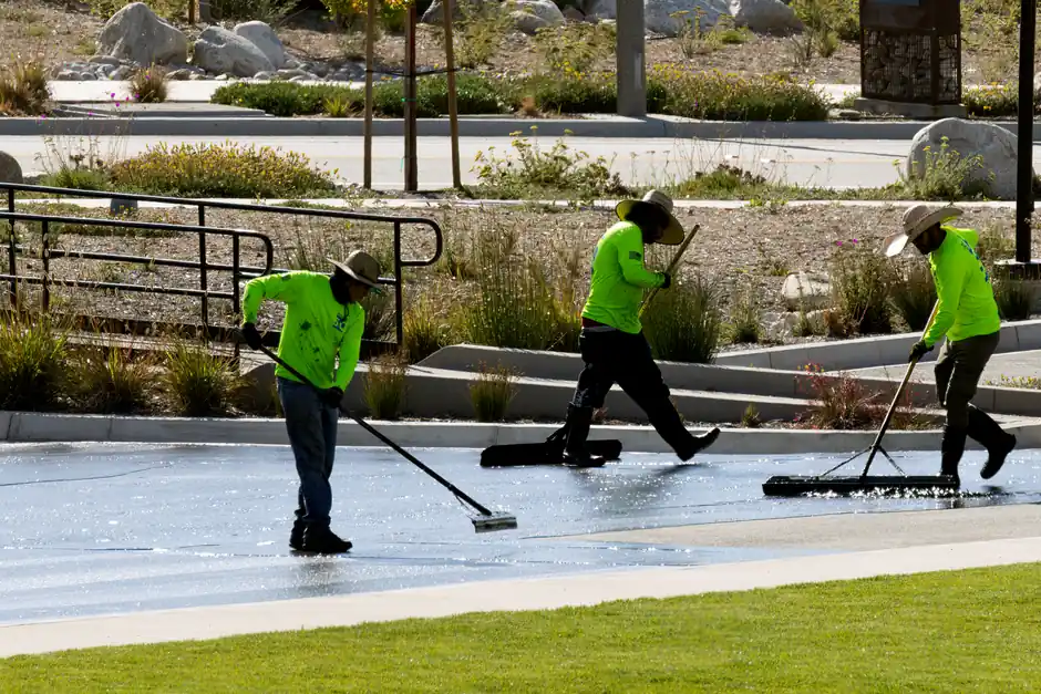 Crew of workers in bright uniforms cleaning a park walkway with brooms under sunlight.