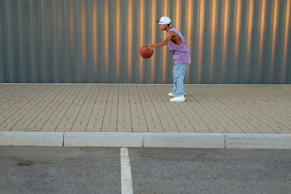 Teenager playing basketball outdoors on a sidewalk, wearing casual attire and cap.